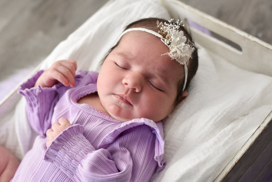 Newborn baby wearing a purple outfit and white headband with flowers.